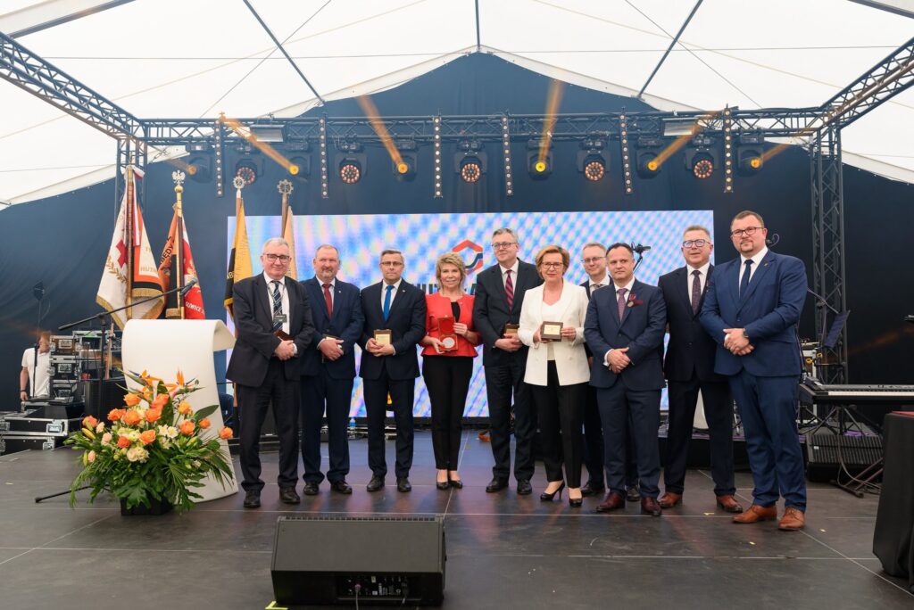 Grupa dziesięciu elegancko ubranych osób stoi na scenie podczas uroczystości pod białym namiotem w tle ekran z logo Huty Łabędy i dekoracyjne oświetlenie Część osób trzyma w dłoniach medale i pamiątkowe tabliczki Z lewej strony podium z mikrofonem oraz bukietem kwiatów obok sztandary firmowe