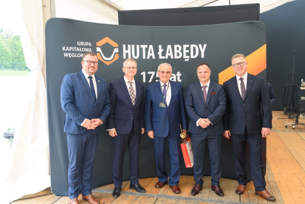 Pięciu mężczyzn w garniturach stoi na drewnianej podłodze przed ciemnym banerem z logo Grupy Kapitałowej Węglokoks i napisem Huta Łabędy 175 lat Jeden z mężczyzn trzyma torbę upominkową i złoty przedmiot w dłoni W tle biały namiot i elementy scenografii eventowej