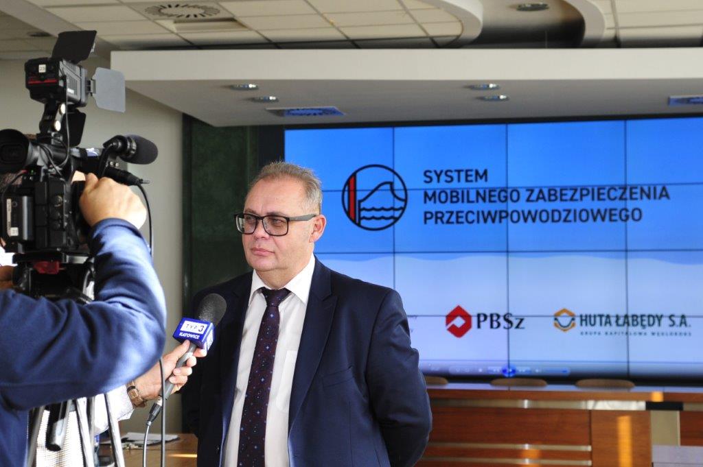 Mężczyzna w garniturze i okularach udziela wywiadu przed kamerą TVP Katowice w tle duży ekran z logo i napisem System Mobilnego Zabezpieczenia Przeciwpowodziowego oraz logotypami PBSz i Huta Łabędy S.A. Grupa Kapitałowa Węglokoks Pomieszczenie o nowoczesnym wystroju z sufitowymi panelami oświetleniowymi