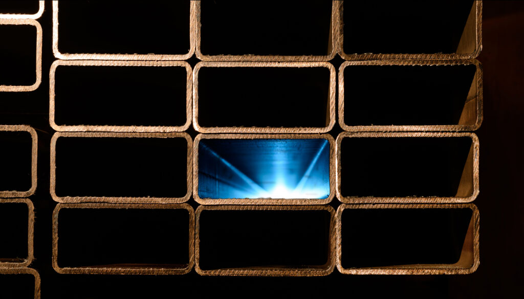Stos ułożonych kształtowników prostokątnych o metalicznej strukturze z widocznymi krawędziami cięcia Jeden z profili oświetlony niebieskim światłem tworzącym efekt głębi i kontrastu na tle ciemnych wnętrz pozostałych kształtowników